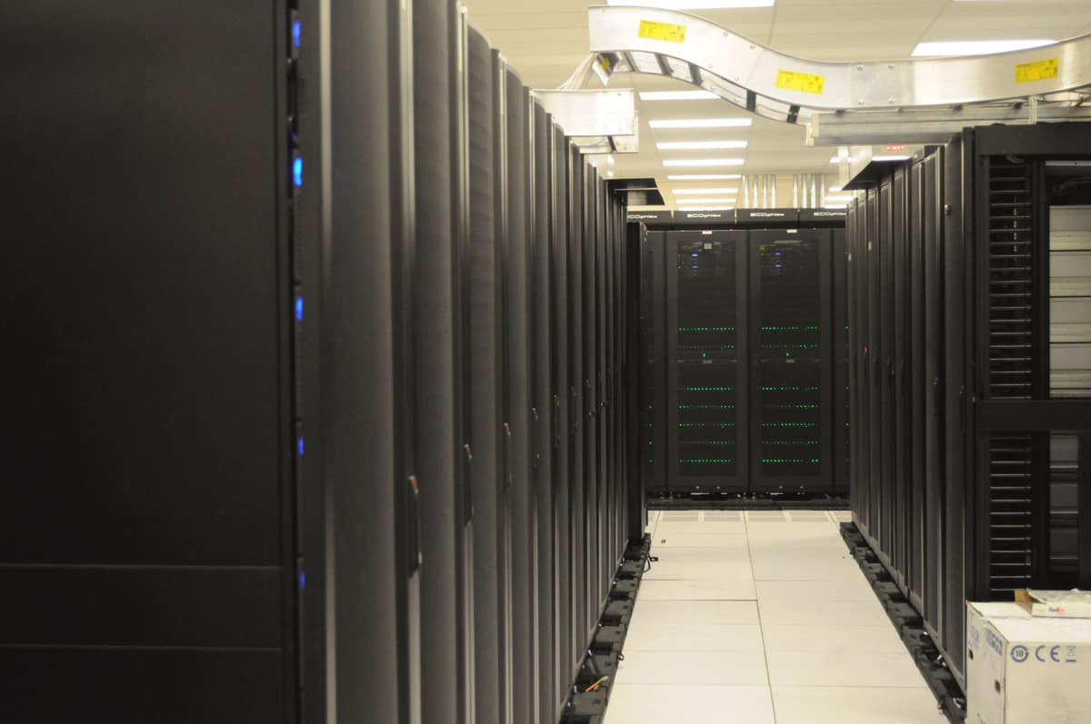 Long aisle between rows of black server racks in a data centre, with cable trays overhead and a raised floor