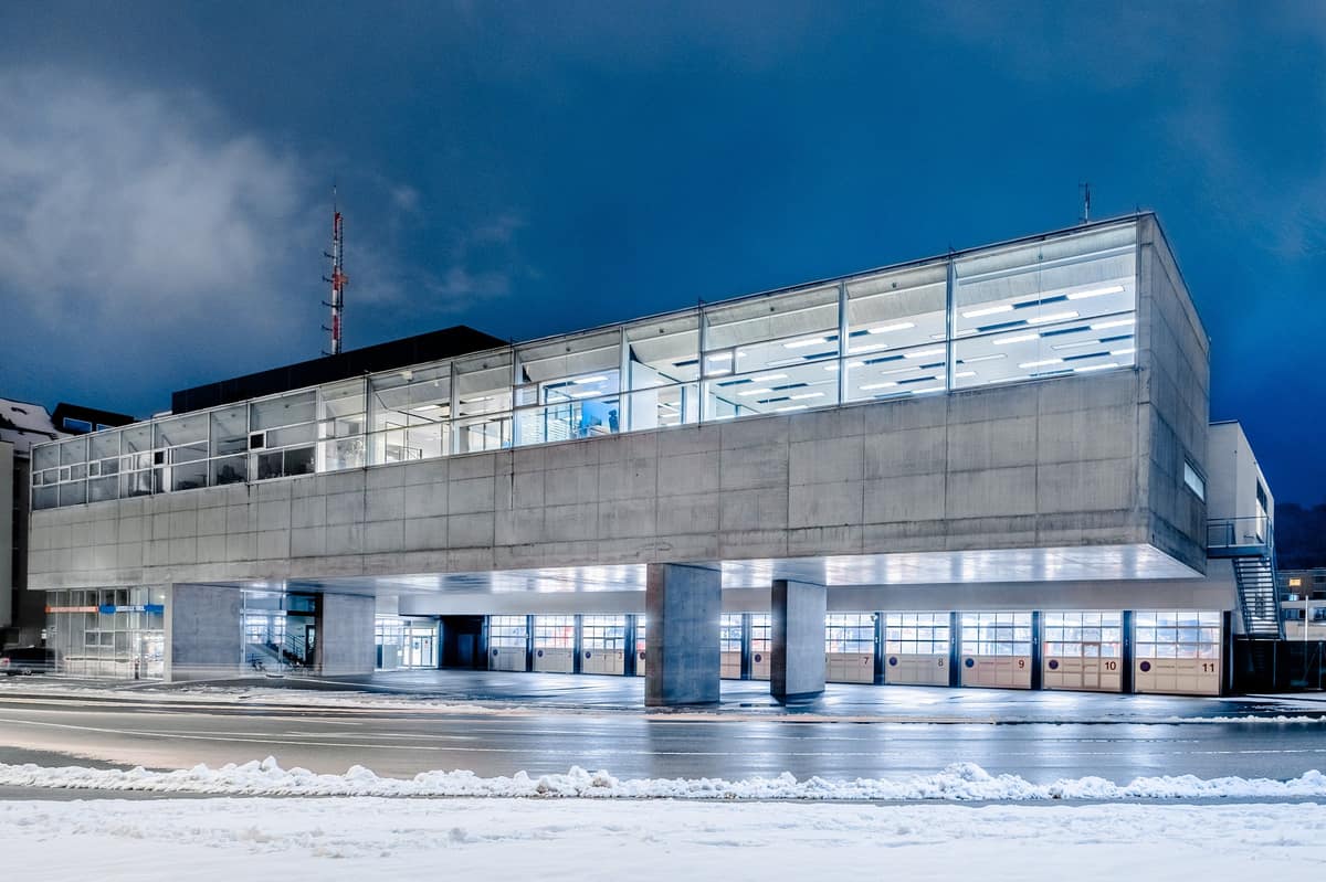 A modern public-sector emergency dispatch centre building at dusk, its glazed upper floor lit above a concrete base and a snowy street.