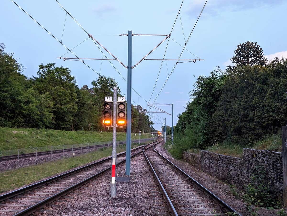 Light-rail signals showing amber aspects on a post beside curving tracks under overhead catenary at dusk.